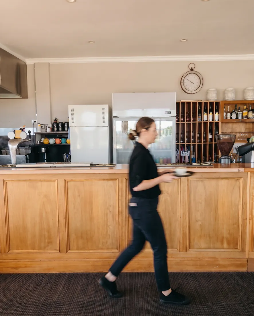 A member of the floor staff in black walking past the timber bar in the Clubhouse Café, caught mid-stride
